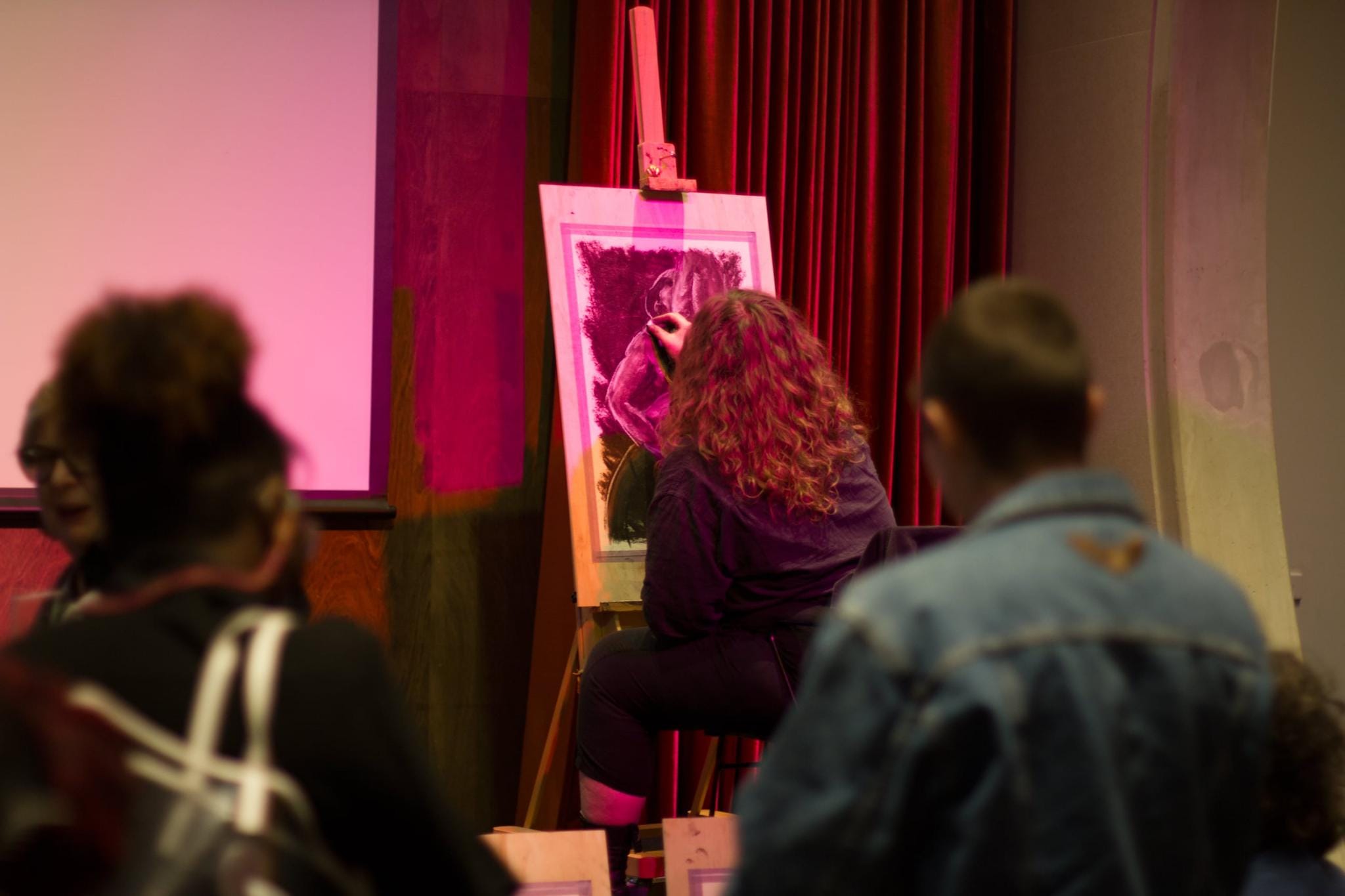 A fat person on stage with back to a watching audience, washed in pink stage lighting. Their mass of light brown curly hair and their light tan body are partially blocking the view of their drawing. They are leaning forward in their seat, drawing a transmasc wheelchair user on paper set up on a board and easel.