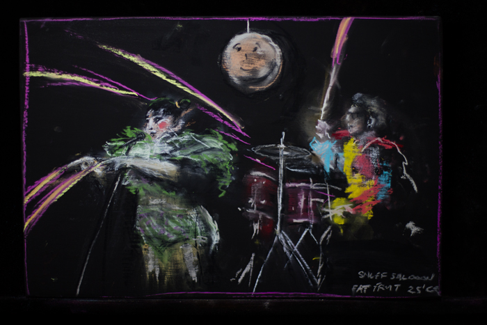 Pastel drawing of Fat Fruit.
Sarah Ward is white, fat and queer, with dark straight hair in two buns. She is wearing a sheer skirt over white and green briefs that highlight the curve of her tummy, and green ruffles around her neck and shoulders. She wears clown-like white makeup, with heavy eye liner and red circle cheeks. She is speaking into a microphone and gesturing toward the crowd with two pointed figures, from which multicoloured laser beams shoot. Behind Sarah is Bec. Bec is sitting at a drum kit, animatedly beating it, one stick in the air from which a laser beam emits. Bec is wearing multicoloured puffy sleeves and top. She is white, queer and has light curly short hair with shaved sides and heavy dark-rimmed glasses.
Above Sarah and Bec is one of the heads they wore during their performance, but it is hanging from the top of the picture like a disco ball. It's is beige with a simplistic face drawn on.