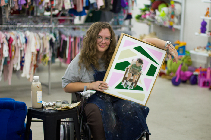Photo of Chris Ferric seated in wheelchair, leaning on their half-finished painting of Jingua Qian. There is a stool with painting supplies and a dirty water bottle next to them. The background is the inside of Savers Footscray, with a rack of clothing and shelf of bric-a-brac